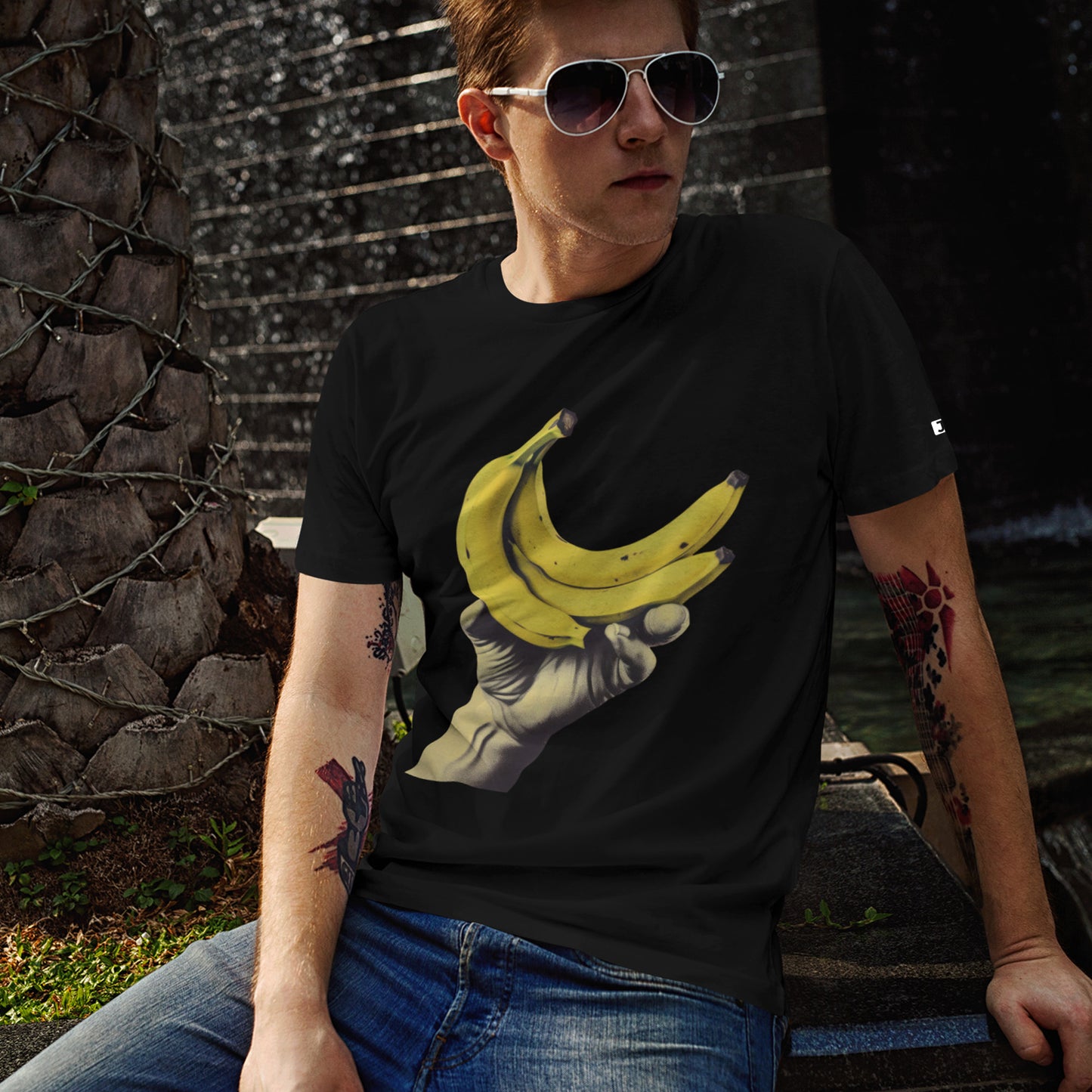 Unisex "Estado Banenero" T-Shirt - Male model outdoors, sitting, wearing sunglasses, in black color.